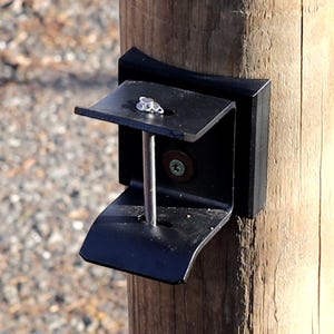 May include: A black metal bracket attached to a wooden post. The bracket has a U-shaped design with a flat top and bottom, and a vertical support rod. A screw is visible on the top surface. The bracket appears to be for mounting or support.