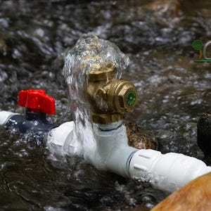 May include: A close-up of a water system with a red valve, brass fitting, and white pipes. Water flows over the brass fitting. The system is partially submerged in a stream with rocks.