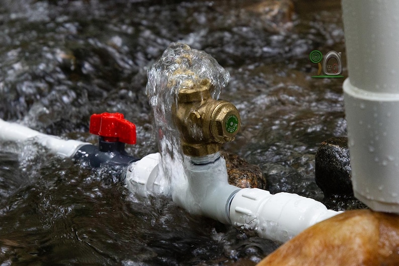 May include: A close-up of a water system with a red valve, brass fitting, and white pipes. Water flows over the brass fitting. The system is partially submerged in a stream with rocks.
