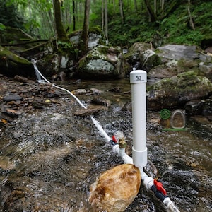 May include: A white PVC pipe with the number 30 printed on top is positioned in a flowing stream. Red and blue valves are attached to the pipe, with water flowing out. The background features a forest with rocks and greenery.