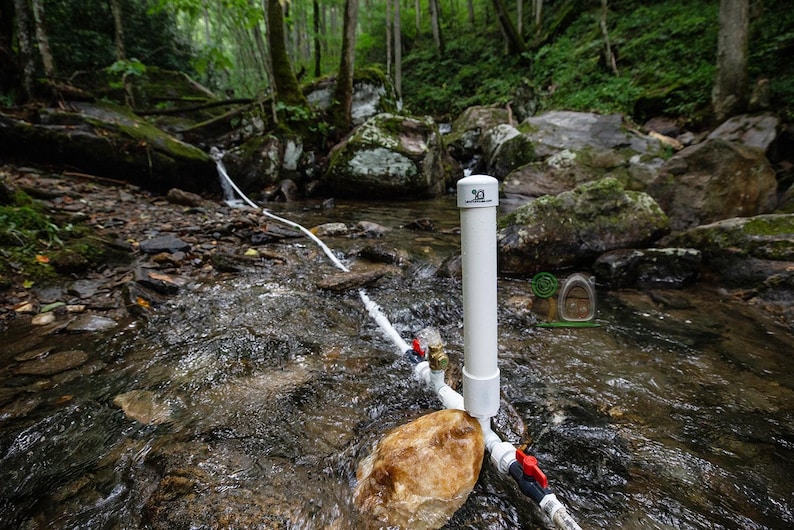 May include: A white PVC pipe with the number 30 printed on top is positioned in a flowing stream. Red and blue valves are attached to the pipe, with water flowing out. The background features a forest with rocks and greenery.