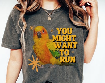 Könnte beinhalten: Dunkelgraues T-Shirt mit einem gelb-grünen Papageien-Motiv. Der Text "YOU MIGHT WANT TO RUN" ist gelb gedruckt. Das T-Shirt hat einen Vintage-Look.