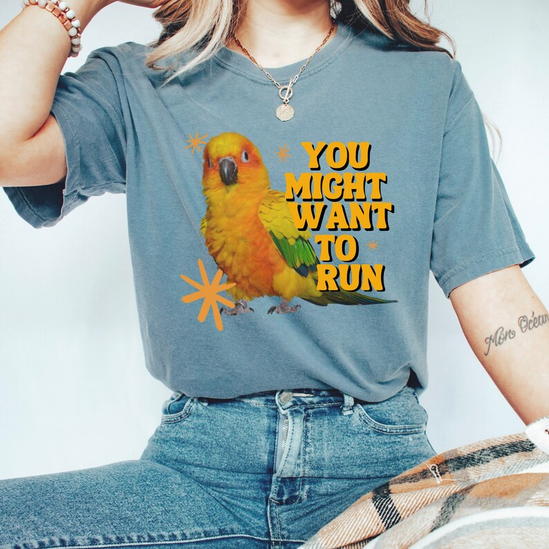 May include: A blue-grey t-shirt featuring a vibrant illustration of a yellow and orange parrot with green accents. The text "YOU MIGHT WANT TO RUN" is printed in yellow and black. The shirt is made of a soft material.