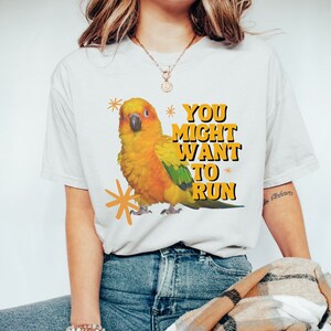 May include: White t-shirt featuring a vibrant illustration of a yellow, orange, and green parrot with the text "YOU MIGHT WANT TO RUN" in orange and black. The shirt is paired with blue jeans.