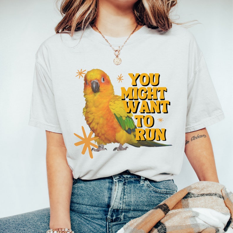 May include: White t-shirt featuring a vibrant illustration of a yellow, orange, and green parrot with the text "YOU MIGHT WANT TO RUN" in orange and black. The shirt is paired with blue jeans.