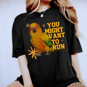 May include: Black t-shirt featuring a vibrant illustration of a yellow, orange, and green parrot with the text "YOU MIGHT WANT TO RUN" in yellow. The design includes orange starburst accents, creating a playful and eye-catching graphic.