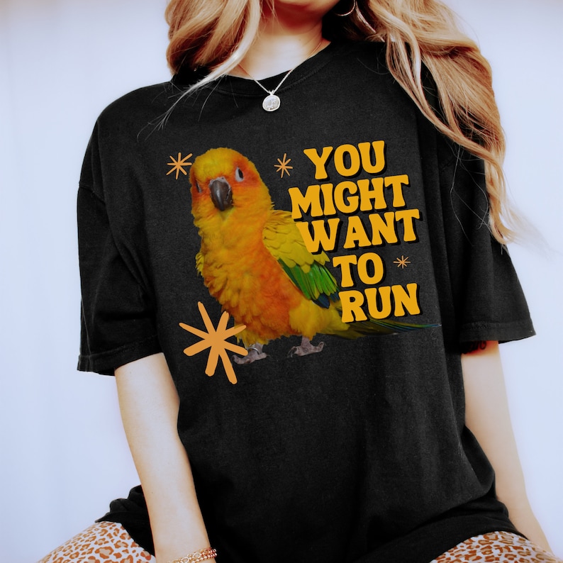 May include: Black t-shirt featuring a vibrant illustration of a yellow, orange, and green parrot with the text "YOU MIGHT WANT TO RUN" in yellow. The design includes orange starburst accents, creating a playful and eye-catching graphic.