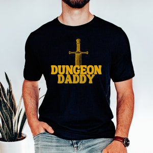 May include: A black t-shirt with a gold sword graphic and the text "Dungeon Daddy" printed on the front.