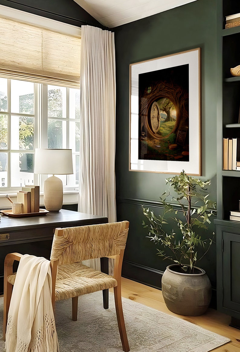 May include: A home office with a dark green wall, a wooden desk, a woven chair, a white lamp, and a framed print of a door in a tree. The print is in a dark brown frame and is hanging on the wall. There is a small potted plant in the corner of the room.