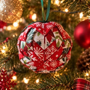 May include: A spherical fabric ornament featuring a folded patchwork design in red, white, and green. The fabric displays patterns of snowflakes, poinsettias, and foliage. A thin green ribbon is attached to the top for hanging on a tree.