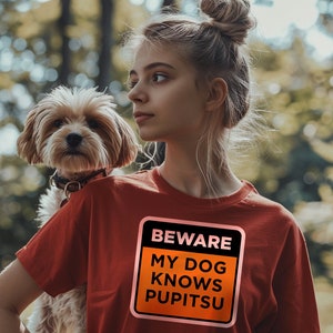 May include: A woman with blonde hair in a bun is wearing a red t-shirt with a black and orange sign that says "BEWARE MY DOG KNOWS PUPITSU". She is holding a small brown and white dog in her arms.