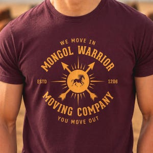Peut inclure: Un t-shirt à manches courtes bordeaux avec un motif graphique doré représentant une silhouette de cheval centrale, des flèches rayonnantes et le texte : WE MOVE IN MONGOL WARRIOR MOVING COMPANY ESTD 1206 YOU MOVE OUT en cercle.