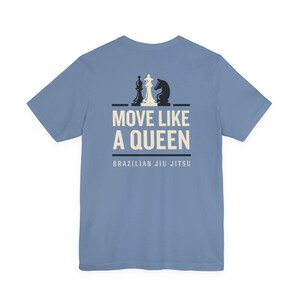May include: A dusty blue short-sleeve t-shirt featuring a graphic on the back. The design includes a white chess queen flanked by two black chess pieces, with the text MOVE LIKE A QUEEN and BRAZILIAN JIU-JITSU printed in white below.