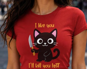 May include: A red short-sleeved t-shirt featuring a graphic of a black cat holding a silver knife. Above the cat, the text reads I like you, and below the cat, the text reads I'll kill you last. The cat has large white eyes and pink ears.