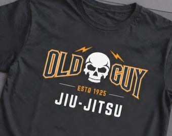 Op de afbeelding: Een zwart t-shirt met korte mouwen en een grafische print van een witte schedel, oranje bliksemschichten en de tekst "OLD GUY" in oranje en zwart. Onder de schedel staat de tekst "ESTD 1925" en "JIU-JITSU" in witte hoofdletters.