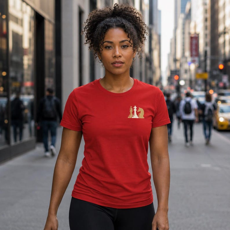 May include: A red short-sleeved t-shirt featuring a small gold graphic on the left chest area. The graphic depicts a stylized chess king and knight piece. The shirt has a crew neckline and a standard fit, constructed from a solid red fabric.