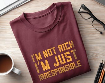 May include: Burgundy crew neck t-shirt with the text "I'M NOT RICH I'M JUST IRRESPONSIBLE" in a distressed gold font. The shirt is folded on a light wood surface, with a coffee cup, glasses, and pencils nearby.