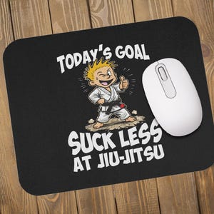 Puede incluir: Alfombrilla de ratón negra con el texto "TODAY'S GOAL SUCK LESS AT JIU-JITSU" y una ilustración de dibujos animados de un niño con uniforme de karate. Un ratón de ordenador blanco está colocado en el lado derecho de la alfombrilla.