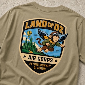 Può includere: Una t-shirt color marrone chiaro con una grafica sul retro. Il design è un emblema a forma di scudo con il testo "LAND OF OZ AIR CORPS FLYING MONKEY DIVISION". Raffigura una scimmia volante con berretto e occhiali da aviatore, un castello verde e una mongolfiera.
