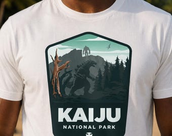 May include: A white t-shirt featuring a graphic print of a shield-shaped emblem. The design depicts a dark silhouette of a large monster, a mountain, pine trees, and a dead tree. The text reads KAIJU NATIONAL PARK in white capital letters.