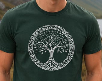 May include: Dark green t-shirt featuring a white Celtic tree of life design. The tree is within a circular frame with intricate knotwork. The shirt is made of a soft material and has short sleeves. The background shows a mountain and lake.
