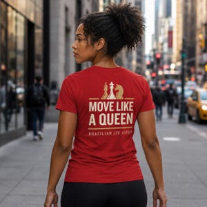 May include: A red short-sleeved t-shirt featuring a graphic on the back with three gold chess pieces above the text "MOVE LIKE A QUEEN" and "BRAZILIAN JIU-JITSU" in a bold, light-colored font. The shirt has a crew neckline and a fitted cut.