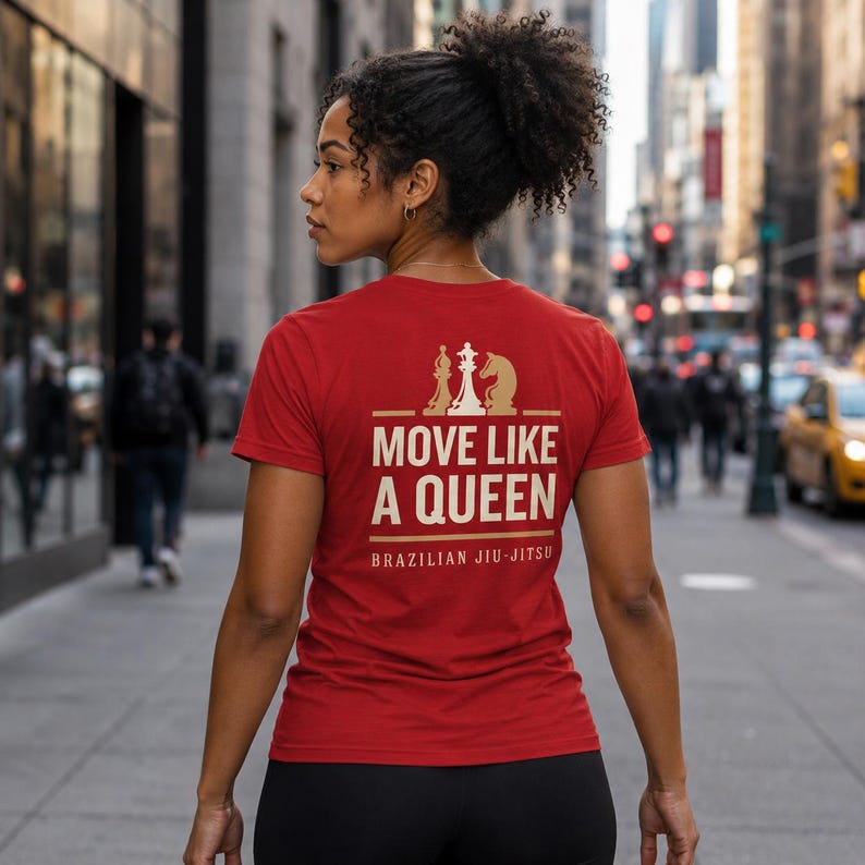 May include: A red short-sleeved t-shirt featuring a graphic on the back with three gold chess pieces above the text "MOVE LIKE A QUEEN" and "BRAZILIAN JIU-JITSU" in a bold, light-colored font. The shirt has a crew neckline and a fitted cut.