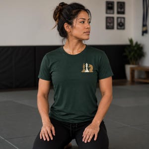 May include: A dark green short-sleeved t-shirt featuring a small graphic on the left chest area. The graphic depicts three chess pieces in a row, including a white queen in the center flanked by two tan-colored knight chess pieces.