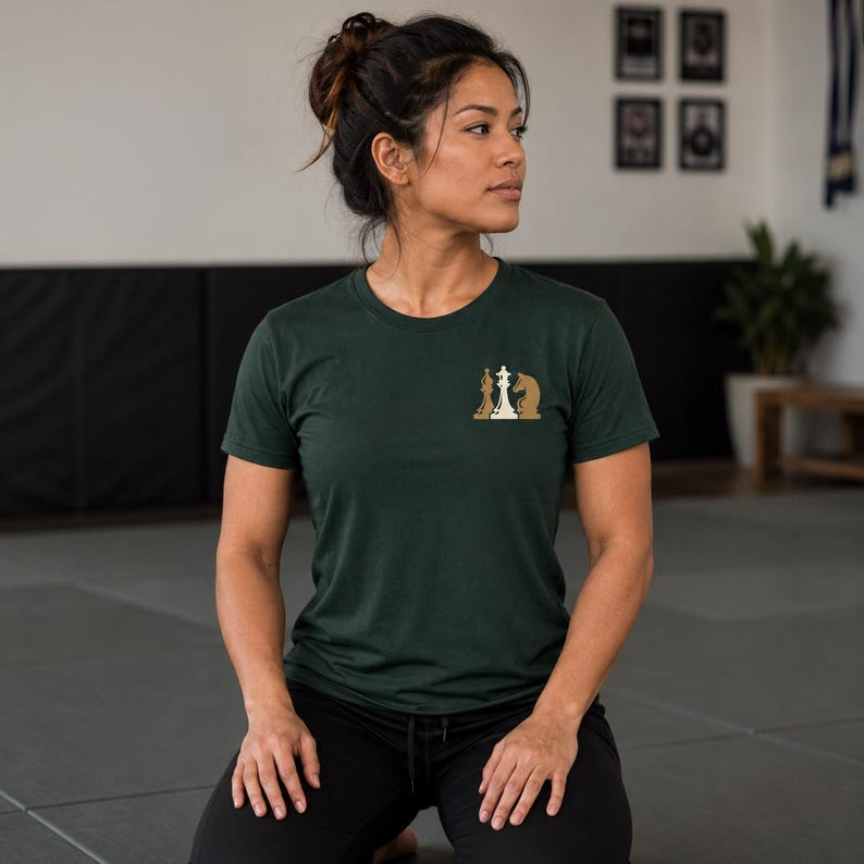 May include: A dark green short-sleeved t-shirt featuring a small graphic on the left chest area. The graphic depicts three chess pieces in a row, including a white queen in the center flanked by two tan-colored knight chess pieces.