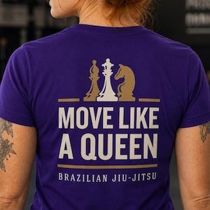 May include: A purple short-sleeved t-shirt featuring a graphic on the back. The design includes gold and white chess pieces above the text MOVE LIKE A QUEEN in large white letters, with the words BRAZILIAN JIU-JITSU printed in smaller white text below.