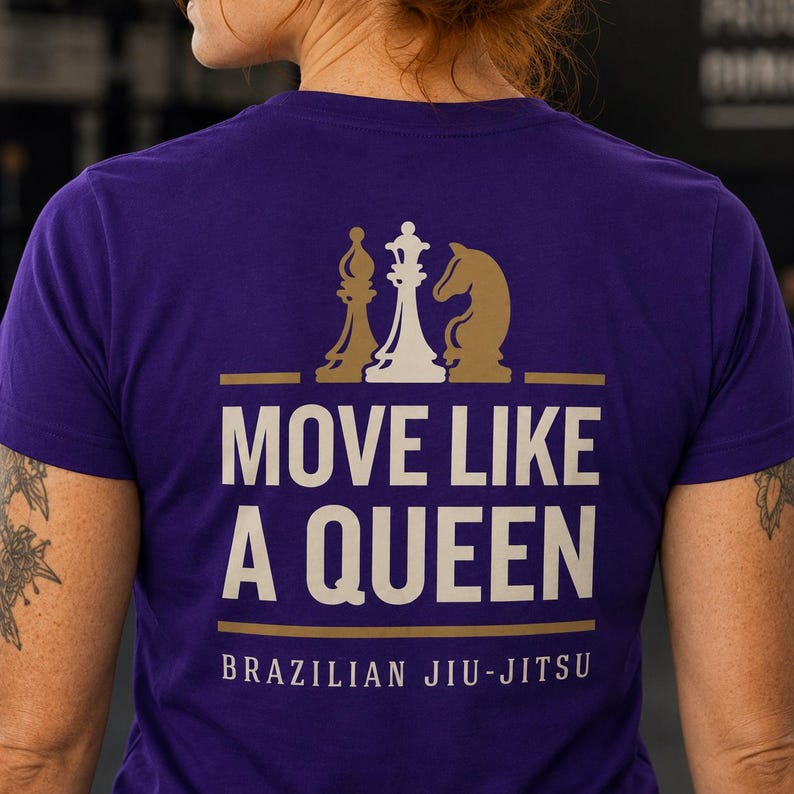 May include: A purple short-sleeved t-shirt featuring a graphic on the back. The design includes gold and white chess pieces above the text MOVE LIKE A QUEEN in large white letters, with the words BRAZILIAN JIU-JITSU printed in smaller white text below.