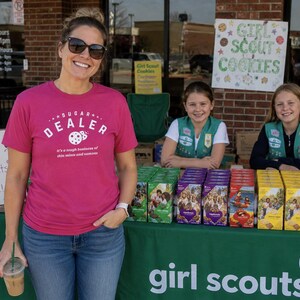 K&ouml;nnte beinhalten: Eine Frau in einem pinkfarbenen T-Shirt mit der Aufschrift "SUGAR DEALER" steht vor einem Girl Scout Cookie-Stand. Zwei junge M&auml;dchen in Girl Scout-Uniformen stehen hinter dem Tisch, der mit Keksschachteln bedeckt ist.