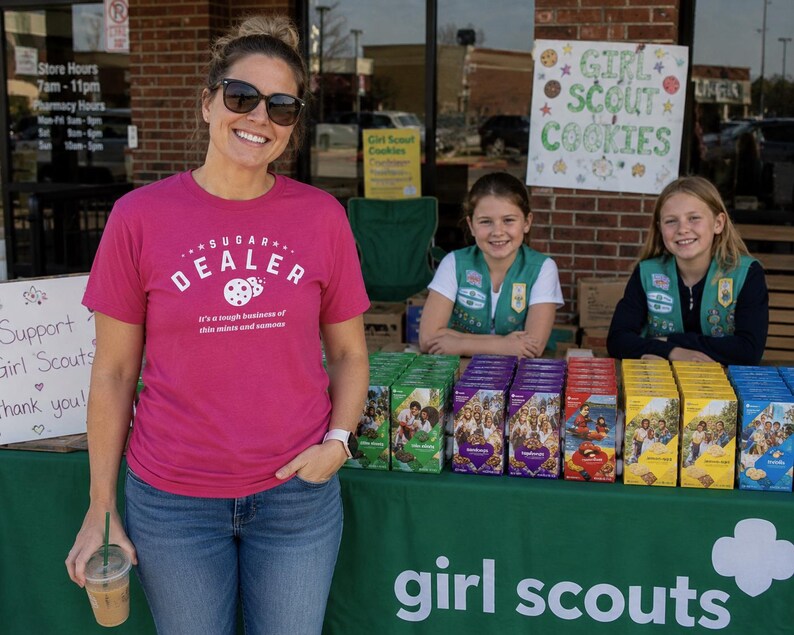 K&ouml;nnte beinhalten: Eine Frau in einem pinkfarbenen T-Shirt mit der Aufschrift "SUGAR DEALER" steht vor einem Girl Scout Cookie-Stand. Zwei junge M&auml;dchen in Girl Scout-Uniformen stehen hinter dem Tisch, der mit Keksschachteln bedeckt ist.