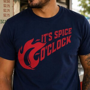 Könnte beinhalten: Marineblaues T-Shirt mit rotem Aufdruck "IT'S SPICE O'CLOCK" und einer stilisierten Flammen-Grafik. Das Design befindet sich auf der Vorderseite. Das T-Shirt besteht aus einem weichen Material.