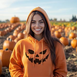 May include: An orange hooded sweatshirt with a jack-o'-lantern face design. The person is smiling in a pumpkin patch with many orange pumpkins in the background. The sky is blue and the field is brown.