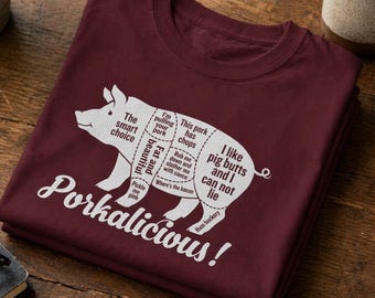 Könnte beinhalten: Ein weinrotes T-Shirt mit einer weißen Grafik eines Schweins, das mit Fleischstücken und humorvollen Sätzen beschriftet ist. Der Text "Porkalicious!" steht unter dem Schwein. Das T-Shirt liegt gefaltet auf einer Holzoberfläche.