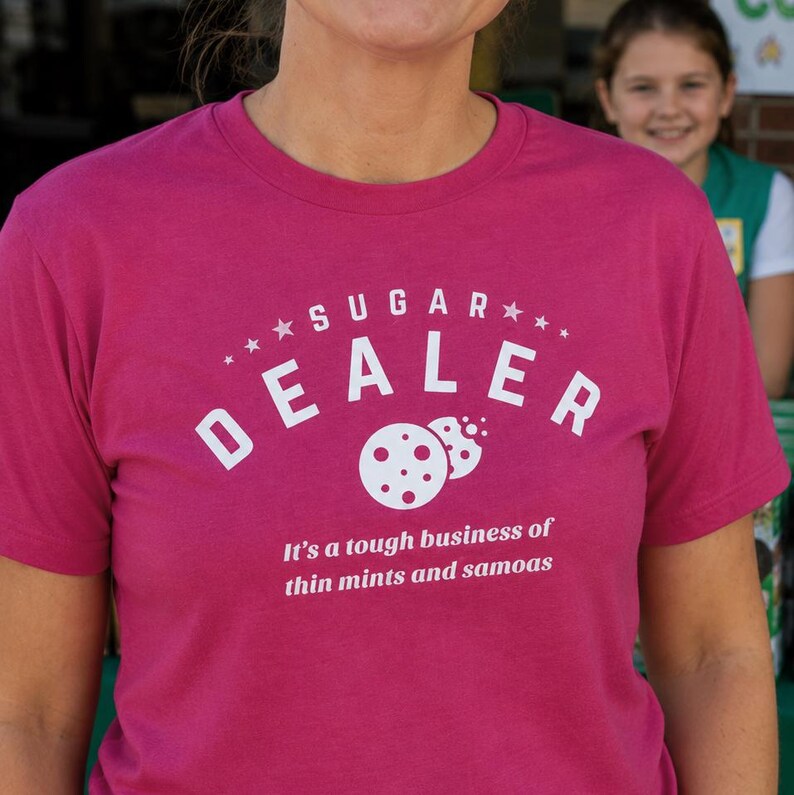 K&ouml;nnte beinhalten: Ein magentafarbenes T-Shirt mit dem wei&szlig;en Schriftzug "SUGAR DEALER" &uuml;ber einer Keksgrafik. Darunter steht der Text "It's a tough business of thin mints and samoas".