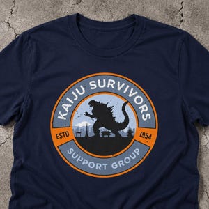 May include: A navy blue t-shirt featuring a circular graphic with an orange border. The design includes a black silhouette of a monster, a mountain, and buildings, with the text KAIJU SURVIVORS SUPPORT GROUP and ESTD 1954 in white and black.