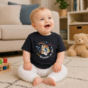 May include: A dark navy blue short-sleeved baby t-shirt featuring a graphic print of a cartoon tiger and a small child grappling. The design includes colourful star and paint splatter accents with the white text "Brazilian jiu-jitsu" printed below.