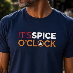 Könnte beinhalten: Ein dunkelblaues T-Shirt mit Rundhalsausschnitt und dem aufgedruckten Text IT'S SPICE O'CLOCK auf der Brust. Das Wort IT'S ist rot, SPICE ist weiß und O'CLOCK ist orange, wobei ein kleines weißes Flammensymbol den Buchstaben O ersetzt.