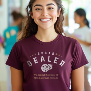 K&ouml;nnte beinhalten: Burgunderfarbenes T-Shirt mit dem wei&szlig;en Schriftzug &bdquo;SUGAR DEALER&ldquo; und einem Keks-Design. Darunter steht der Satz &bdquo;It's a tough business of thin mints and samoas&ldquo;.