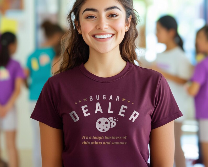 K&ouml;nnte beinhalten: Burgunderfarbenes T-Shirt mit dem wei&szlig;en Schriftzug &bdquo;SUGAR DEALER&ldquo; und einem Keks-Design. Darunter steht der Satz &bdquo;It's a tough business of thin mints and samoas&ldquo;.