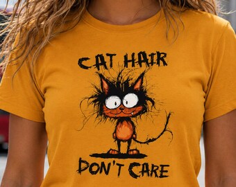 May include: A mustard yellow short-sleeved t-shirt featuring a graphic illustration of a messy-haired orange cat with large white eyes. The text "CAT HAIR" is printed above the cat, and "DON'T CARE" is printed below in a distressed black font.