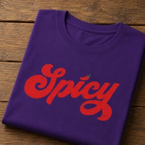 May include: A purple short-sleeved t-shirt featuring the word Spicy printed in a distressed red font across the chest. The letter i is topped with a small green chili pepper icon. The shirt has a crew neckline and a smooth, solid fabric texture.