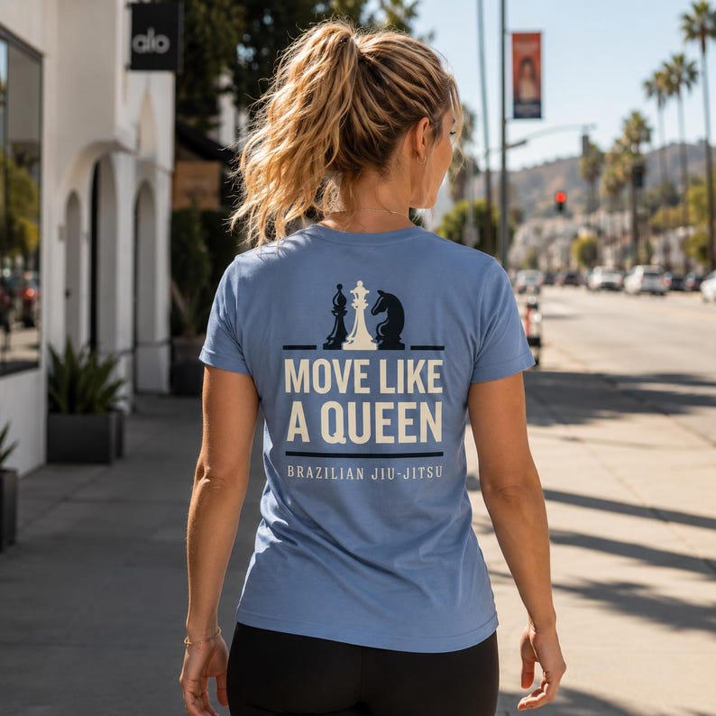 May include: A light blue short-sleeve t-shirt featuring a graphic on the back with three chess pieces above the text "MOVE LIKE A QUEEN" and "BRAZILIAN JIU-JITSU" printed in white and black, with a crew neckline and a standard fit.