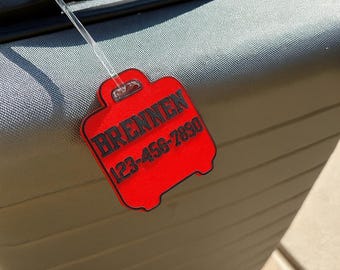 May include: A gray suitcase with a black handle and zipper. A red luggage tag is attached, displaying the name "BRENNEN" and a phone number. The tag is shaped like a suitcase.