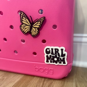 May include: Pink plastic tote bag with a black butterfly and a white sticker that says "Girl Mom" with a black flower in the middle.