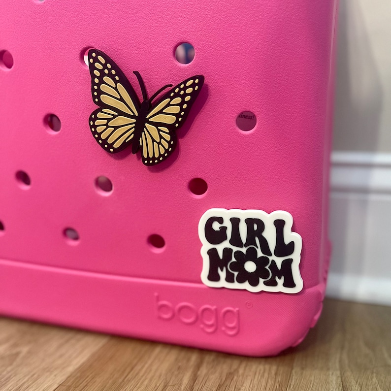 May include: Pink plastic tote bag with a black butterfly and a white sticker that says "Girl Mom" with a black flower in the middle.