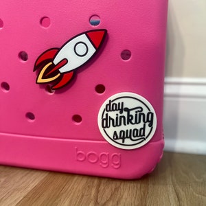 May include: Pink Bogg bag with a white and red rocket and a white circle sticker with the text "day drinking squad" in black.