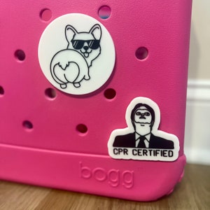 May include: Pink plastic container with a white sticker of a cartoon dog wearing sunglasses and a black sticker of a man with the text "CPR Certified". The container has the word "bogg" printed on the bottom.